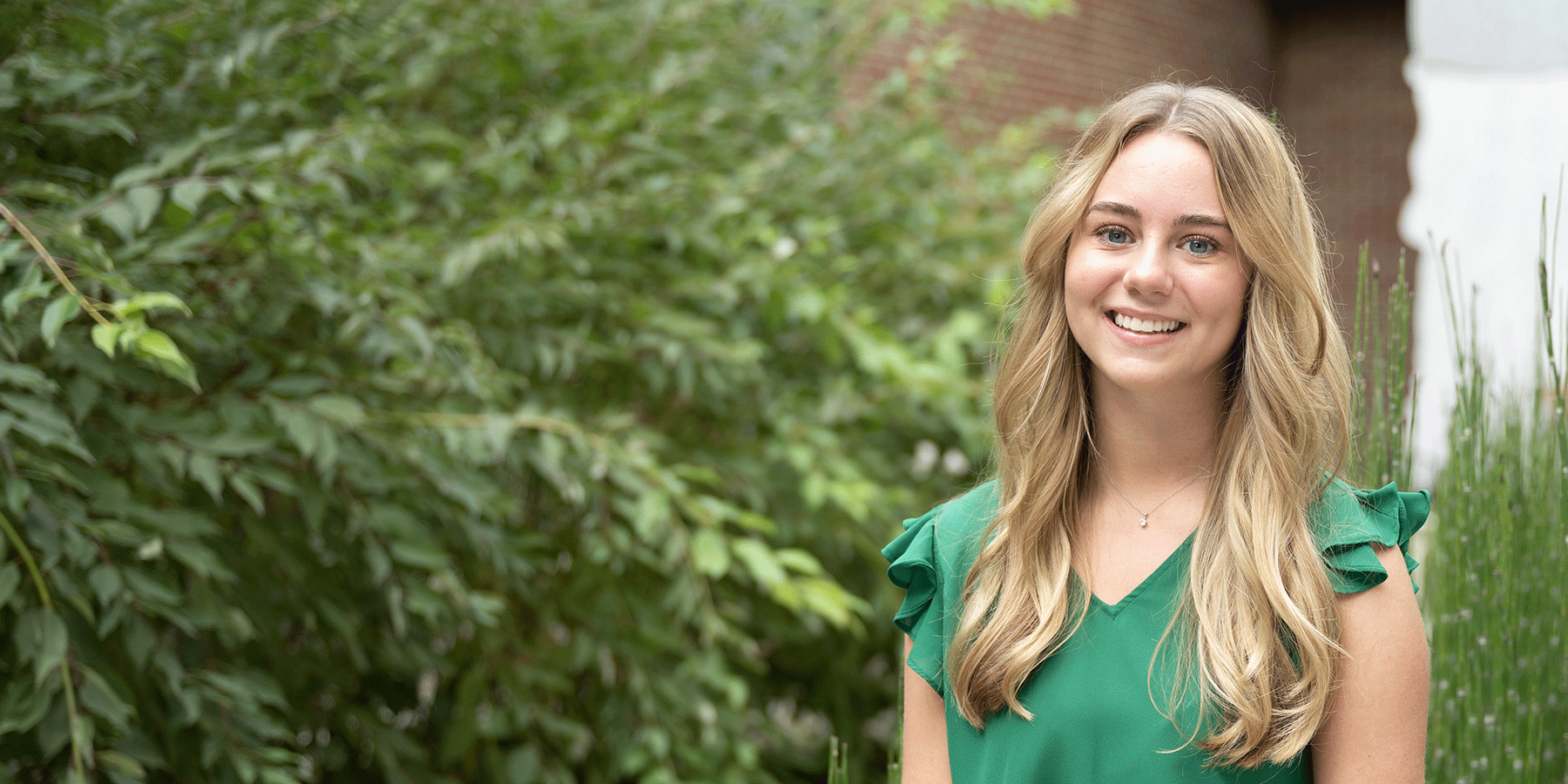 A smiling, blonde, female student wearing a green, shoulder-sleeve blouse is shown outdoors on campus, with green leaves and grasses in the background and to the right of the image.